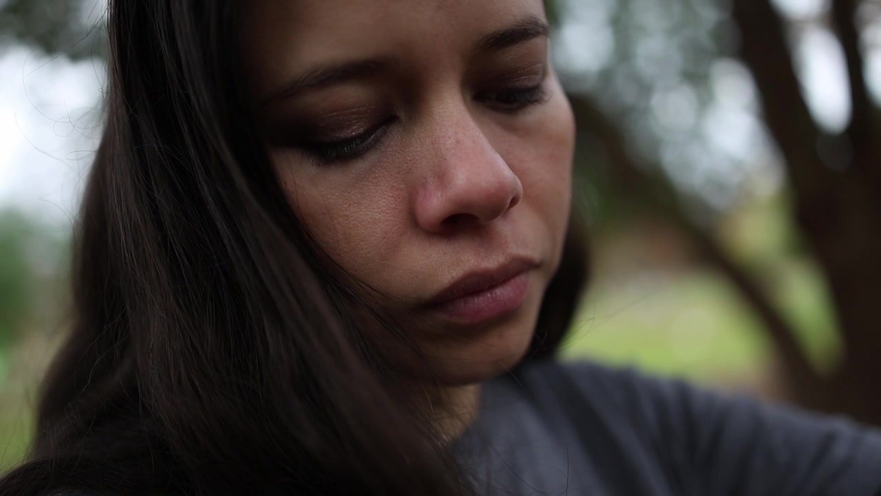 Close-up de uma jovem com cabelo longo e escuro sentada num banco num parque, parecendo imersa em pensamentos. Iluminação natural suave, fundo verde desfocado e foco raso evocam introspecção.