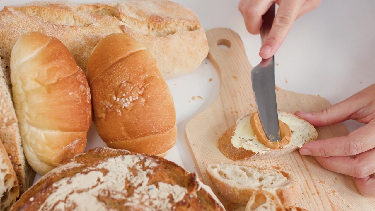 Close-up shot of hands spreading peanut butter on bread amidst various bakery loaves.