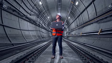 full body of asian male engineer with safety helmet working on a laptop and looking around while standing in underground subway tunnel
