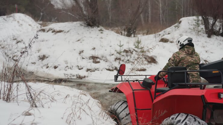 ATV rosso che attraversa la sponda ghiacciata di un ruscello, un pilota con casco regola la marcia e si allinea per il guado, pneumatici che mordono il fango ghiacciato, spruzzi d'acqua bassa, sponde aspre e arbusti senza foglie, controllo preciso e mirato nel freddo.