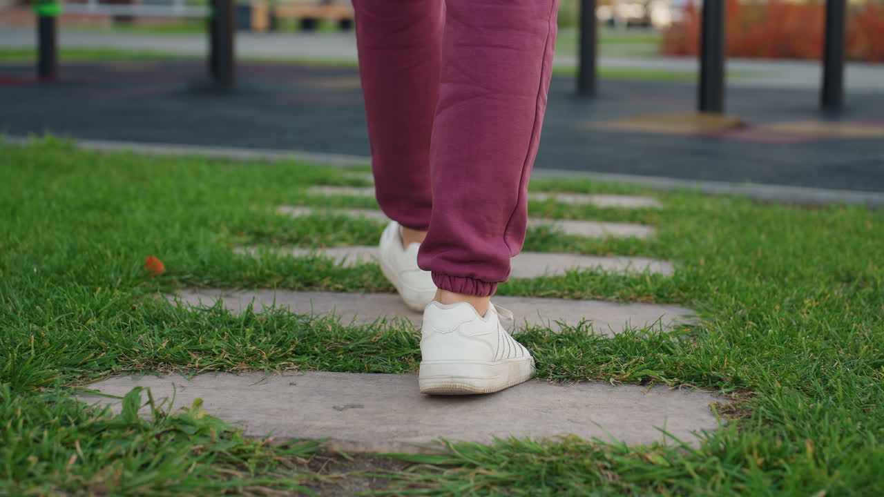 Resilient woman in hoodie carrying sports bag walks on grassy stone path toward exercise bars set within urban outdoor fitness area, showcasing determination and active lifestyle in crisp daylight