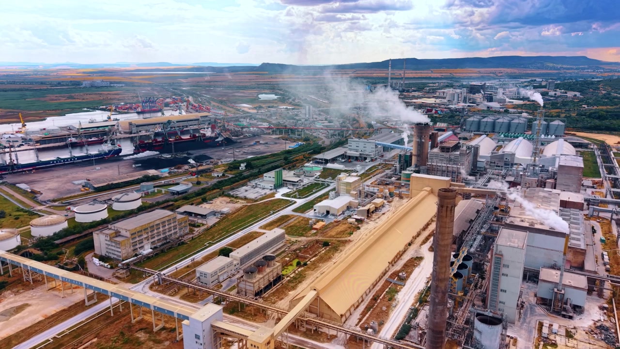 Aerial view of chemical manufacturing plant with smokestacks. Industrial facility emitting smoke near harbor with yellow buildings under cloudy sky.
