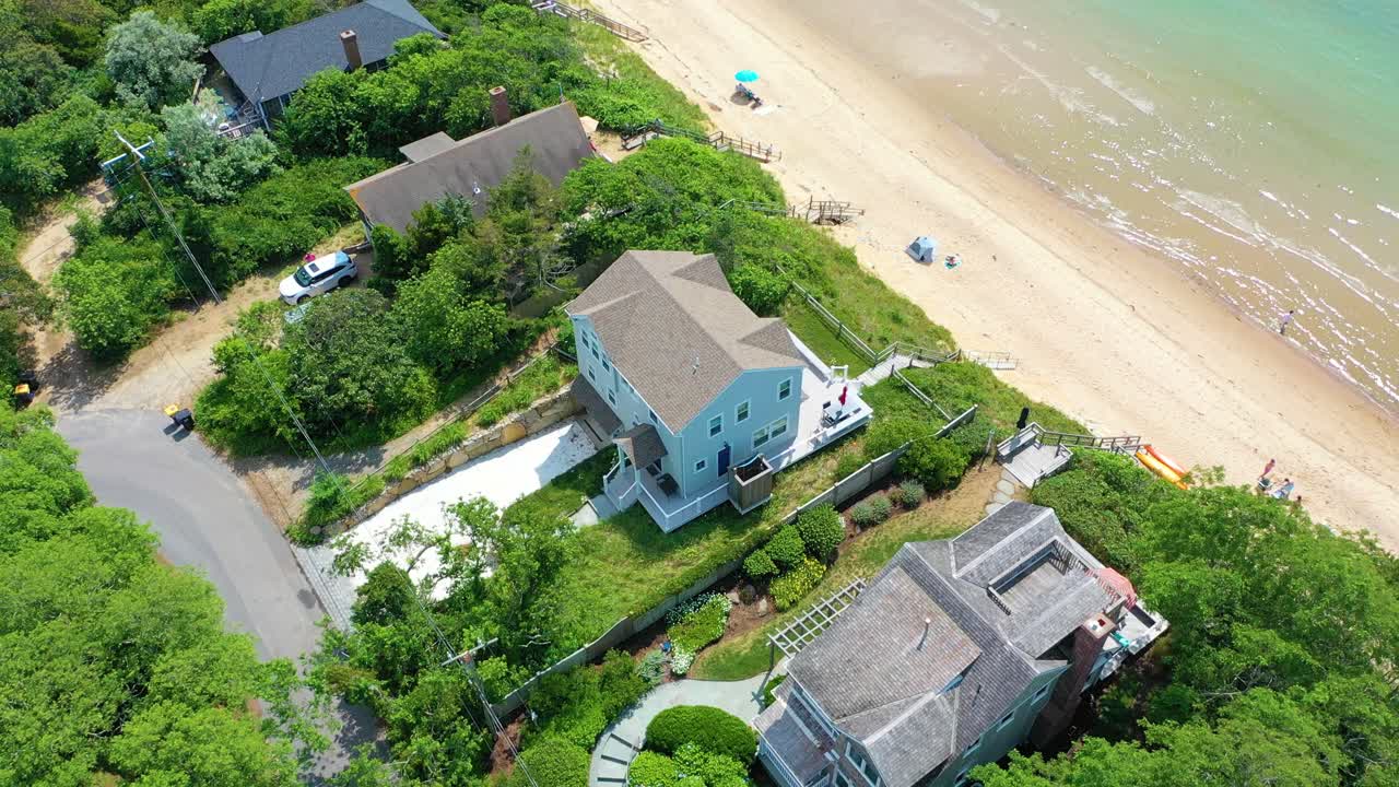 Aerial view of Cape Cod shoreline captures a beachfront home framed by dunes, beachgoers visible below, and turquoise waves glowing under warm summer light