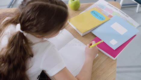 Niña-Sentada-En-El-Escritorio-Y-Escribiendo-En-Un-Cuaderno-Durante-La-Clase-De-Inglés-En-La-Escuela