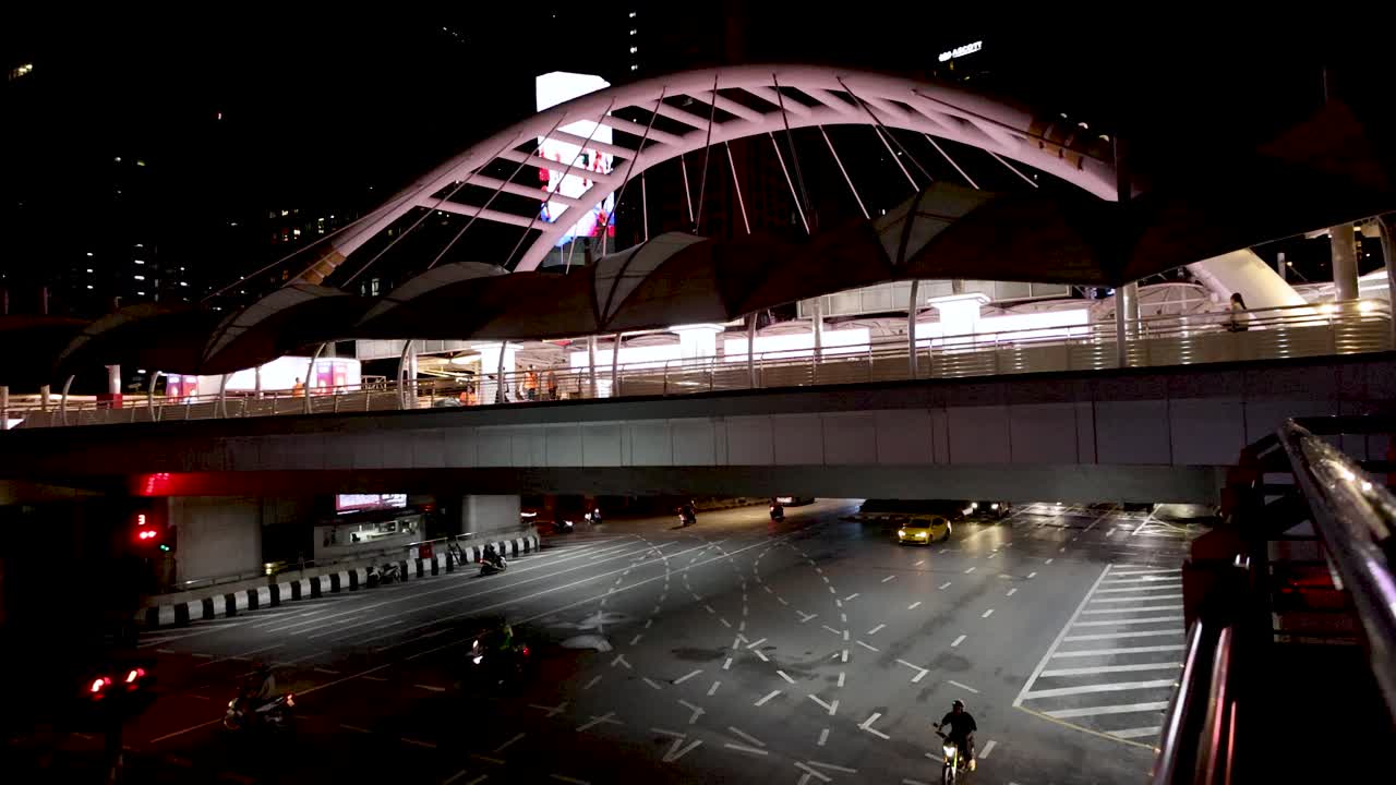 Vehicles and lone pedestrian cross illuminated bridge at night, wide static shot, urban environment