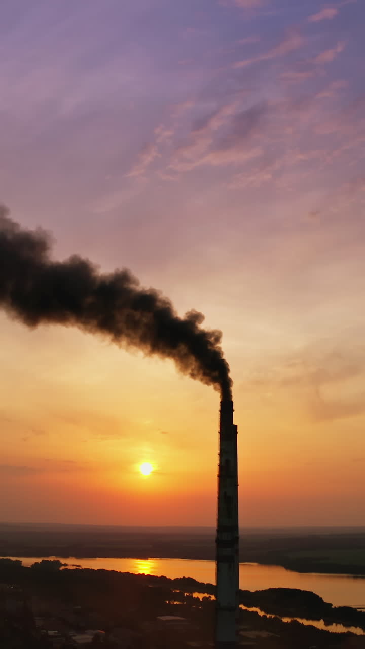 Dark fumes in the atmosphere from industry. Smoking chimney polluting the environment at sunset. Harmful industrial plant near the river in the evening. Vertical video