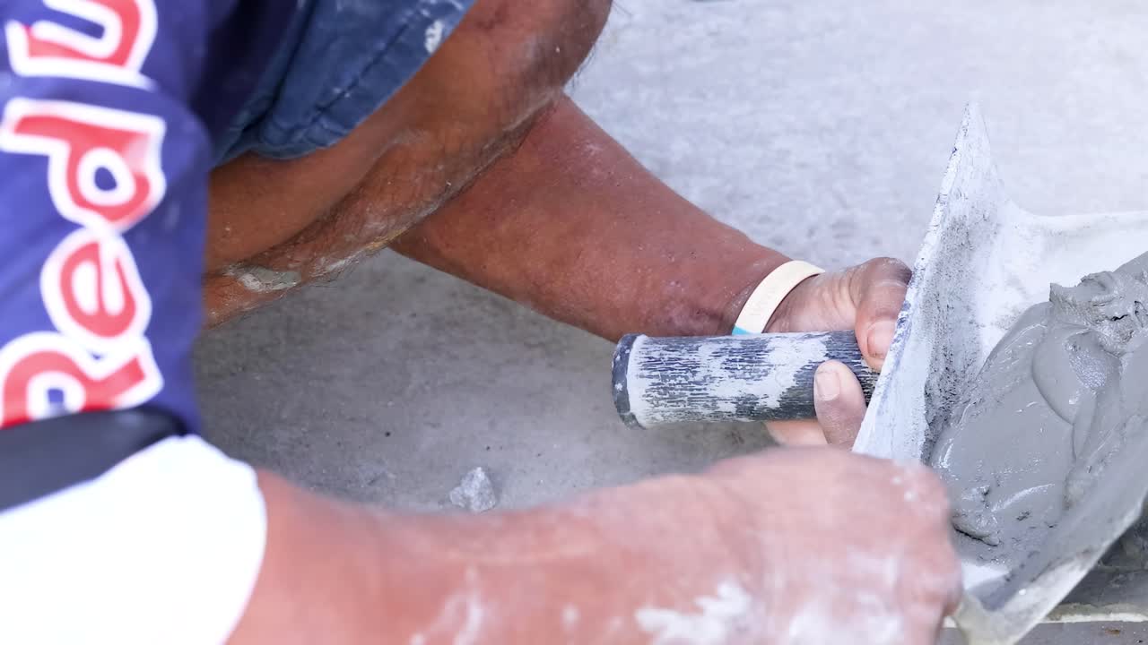 A person spreads cement on a surface outdoors, using a trowel, under bright daylight conditions