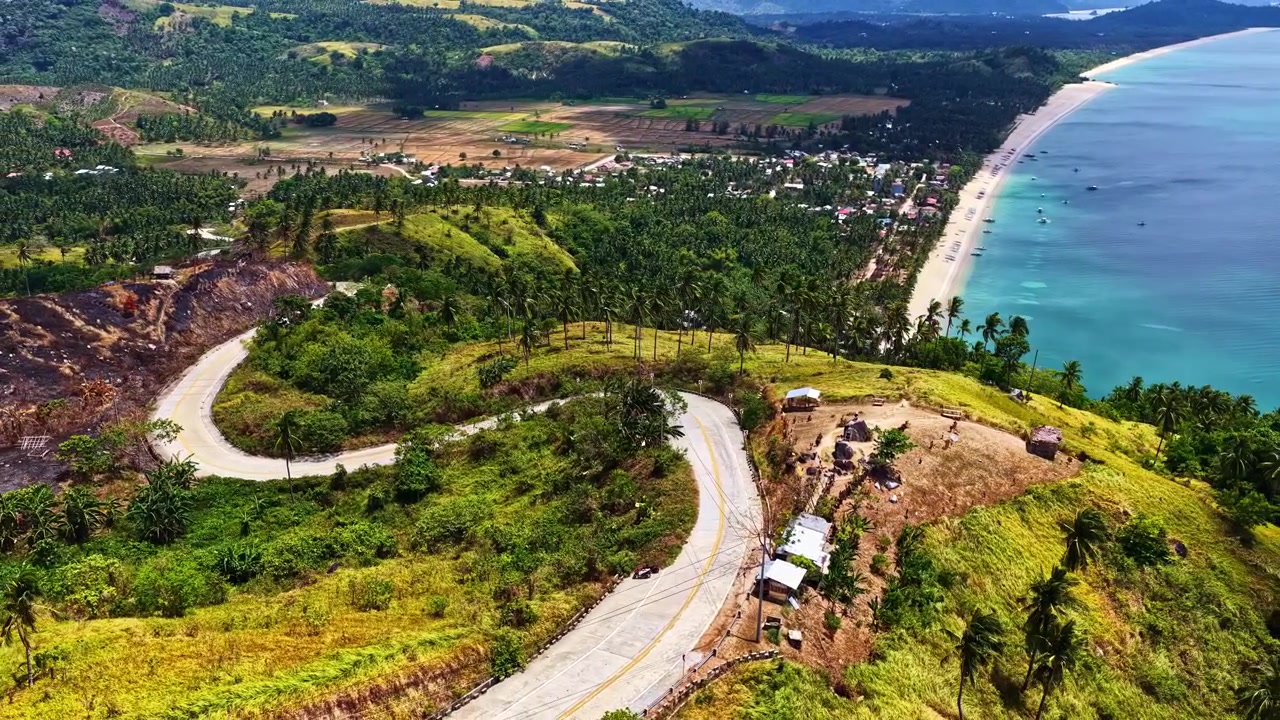 Jalan berkelok-kelok membentang di sepanjang lereng bukit hijau subur menghadap ke laut biru kehijauan di titik pandang San Vicente di Filipina, diambil dari drone