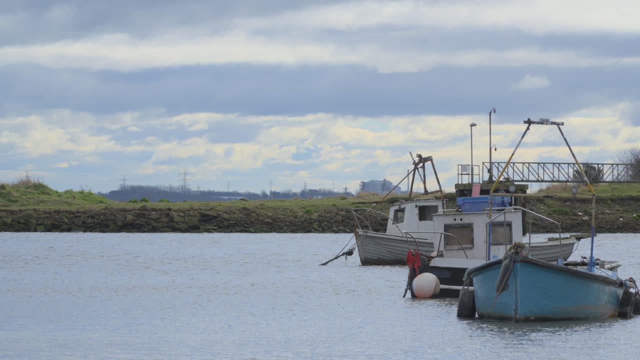 pequeños barcos de pesca que se mueven en la marea que se retira a medida que una nube gruesa se mueve a través del marco con edificios industriales en el horizonte
