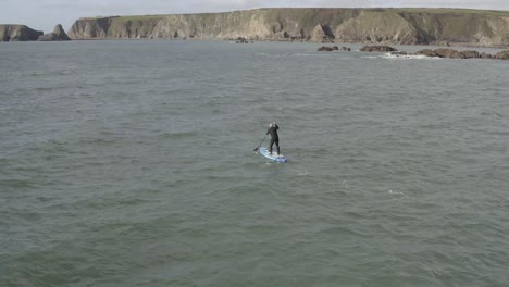 man in wetsuit turns paddle board toward rocky islets and sea cliffs