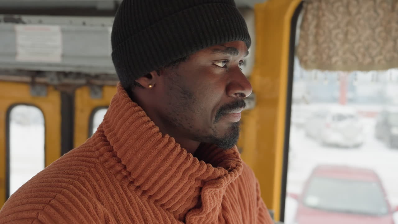 Uomo di colore all'interno di un autobus giallo della città, con maglione lavorato a maglia arancione e berretto nero visibili, mani vicino alla bocca per scaldarsi, sguardo sulla strada innevata, ruoli alternati tra le inquadrature come autista, controllore a bordo