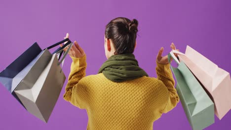 Rear-view-of-woman-holding-coloured-gift-bags-on-purple-background