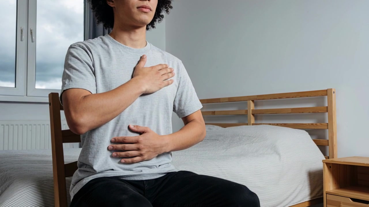 Hombre colocando las manos y cerrando los ojos, practicando respiración profunda en una silla de madera junto a la cama para relajarse