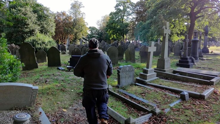 Uomo con cappello e abiti casual che cammina da solo lungo un tranquillo sentiero di cimitero illuminato dal sole