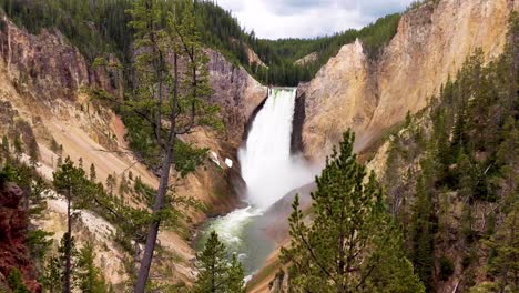 Stunning-static-wide-view-of-huge-waterfalls-and-river-water-flowing-through-rocky-mountain-range,-wind-blowing-gently-against-pine-trees
