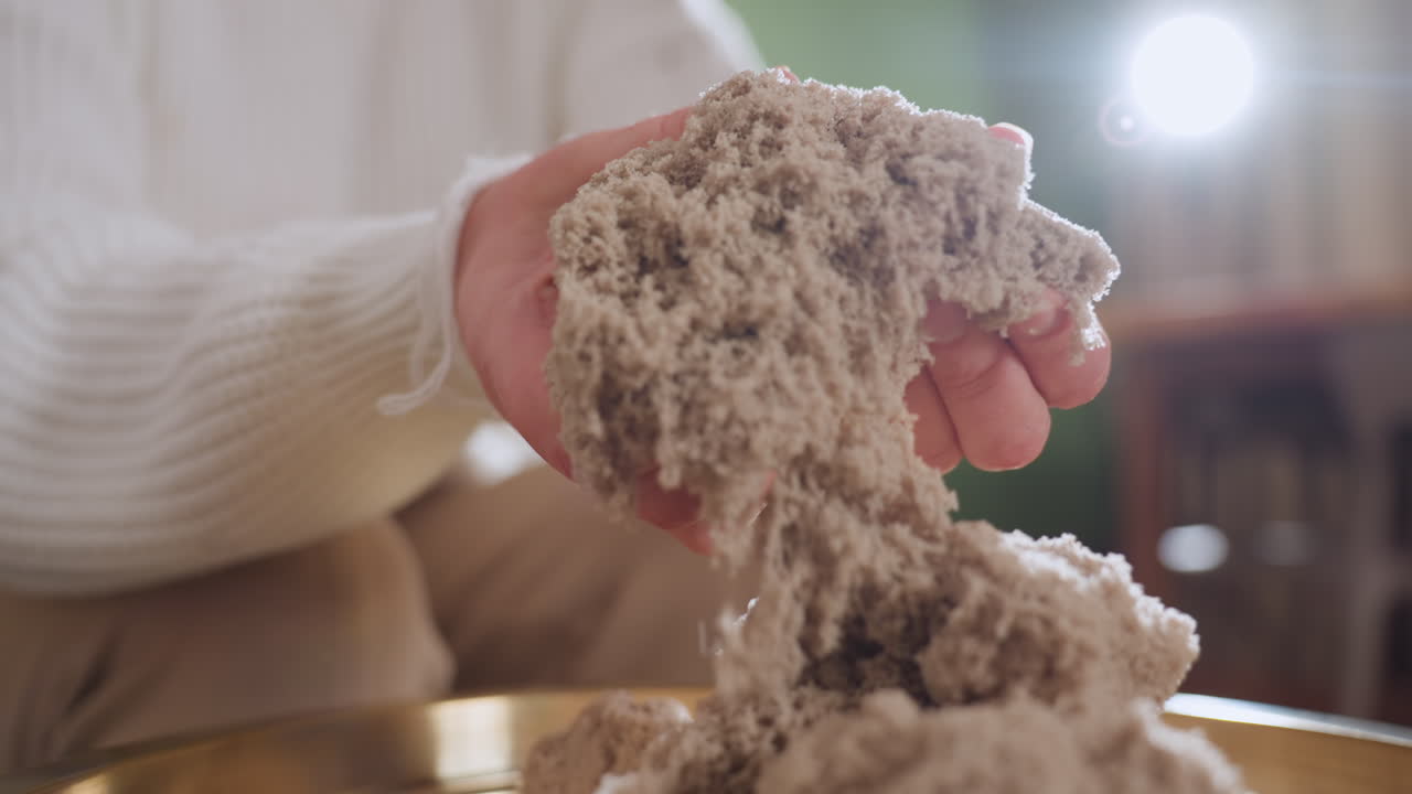 Reflective woman gently lifting soft wet soil mold with both hands during calming sensory therapy session, highlighting tactile texture on golden tray in peaceful indoor space with blurred background
