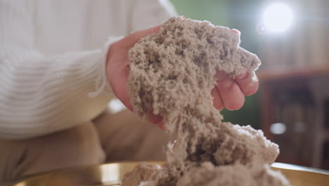 reflective woman gently lifting soft wet soil mold with both hands during calming sensory therapy session, highlighting tactile texture on golden tray in peaceful indoor space with blurred background