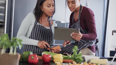 Feliz-Pareja-De-Lesbianas-Birraciales-Preparando-Verduras-Y-Usando-Tabletas-En-La-Cocina,-En-Cámara-Lenta