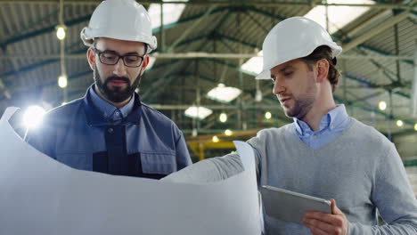 Two-engineer-wearing-helmets-with-tablet-device-and-blueprint-talking-in-a-factory