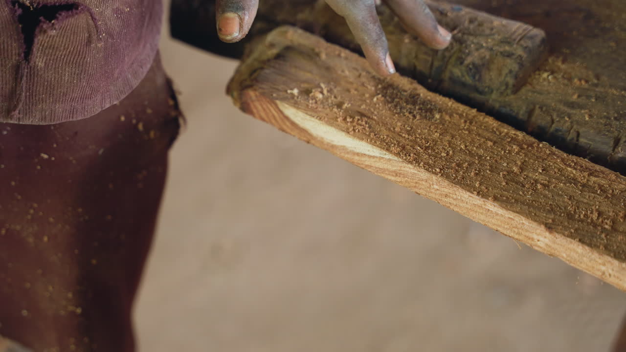 Apprendista che leviga una tavola di legno. Un apprendista falegname nero rifinisce attentamente una superficie di legno testurizzata. Primo piano di mani che levigano con perizia una tavola di legno impolverata in un ambiente di officina rustica.