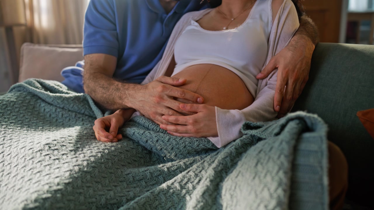 Coppia incinta che si abbraccia sul divano toccandosi la pancia. Primo piano di un momento di legame familiare a casa