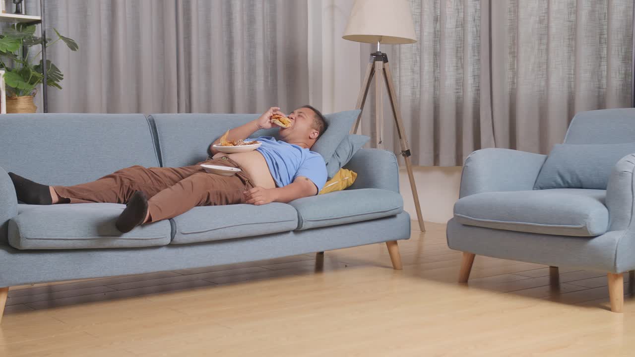 집에서 거실에서 패스트푸드를 먹는 햄버거를 먹고 소파에 누워있는 한 아시아 남자의 몸이 가득 차 있습니다.