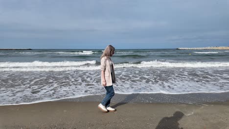 Una-Toma-En-Cámara-Lenta-De-Una-Chica-Rubia-Caminando-Por-La-Playa-Con-Olas-Golpeando-Lentamente-La-Costa