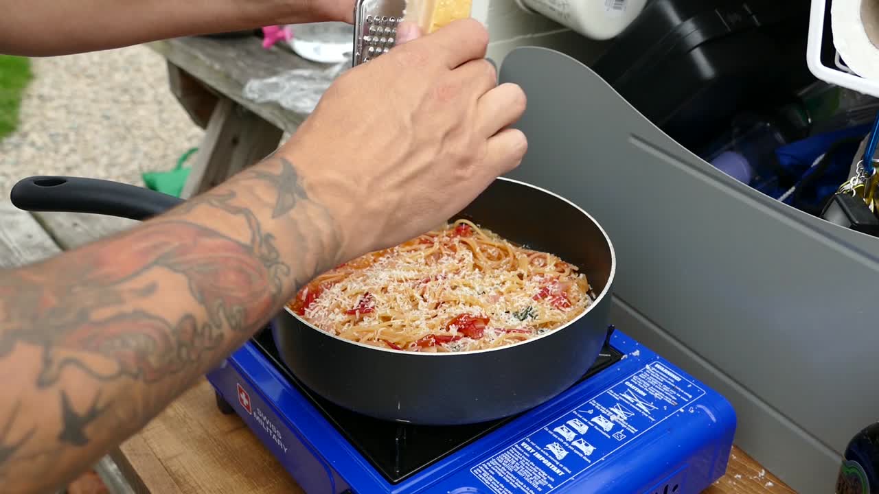 joven con tatuajes en los brazos puso queso parmesano en algunos espaguetis de tomate cocinados en una estufa de camping