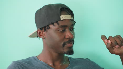 close up of handsome boy wearing cap sitting on red couch, looking off to side at something in background while rubbing his hand against teal wall with calm expression in bright indoor setting