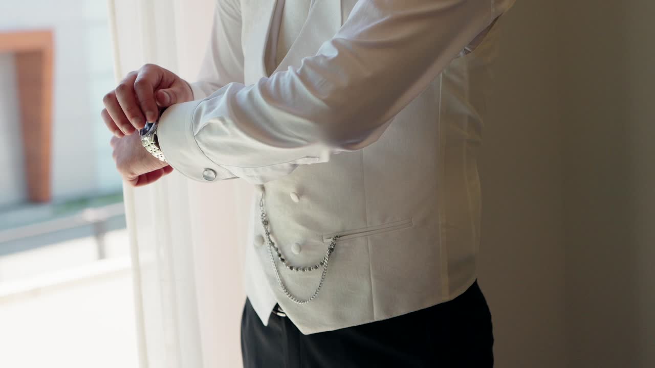 Close up of a groom adjusting a silver chain on a cream waistcoat during wedding preparation