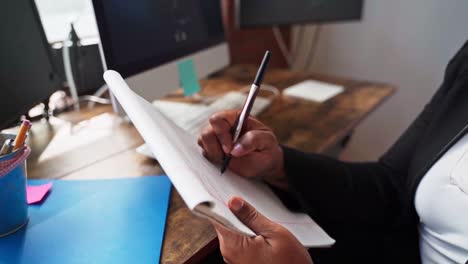 Female-at-home-office-is-working-and-taking-notes-on-a-notepad-while-sitting-next-to-desk-with-computer-screen