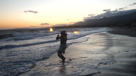 a man fire dancer and performer in silhouette spinning his flaming staff in the ocean water on the beach at sunset in slow motion