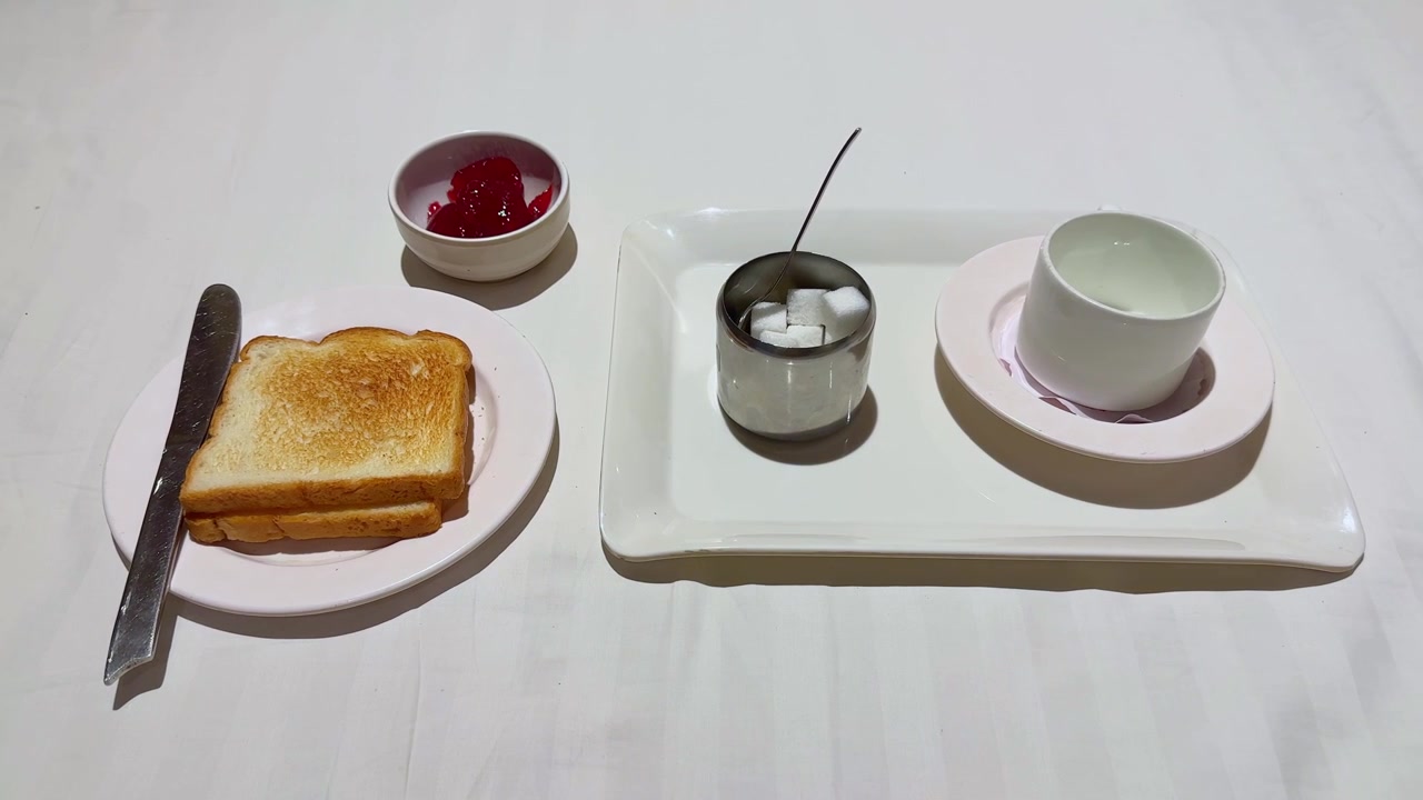 Sakte kamerabevegelse mot en frokosttallerken med ristet brød, smørkniv, syltetøy, sukkerbiter og tekopp arrangert på et rent spisebord
