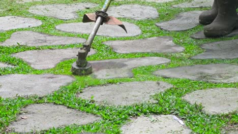 Trabajador-Trabajador-Recortando-La-Hierba-Crecida-Entre-Bloques-De-Hormigón-Usando-Una-Máquina-Cortadora-De-Cepillos-En-El-Camino-Del-Jardín