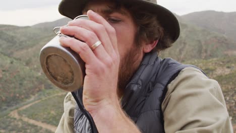 caucasian male survivalist sitting on mountain in wilderness, drinking water and smiling