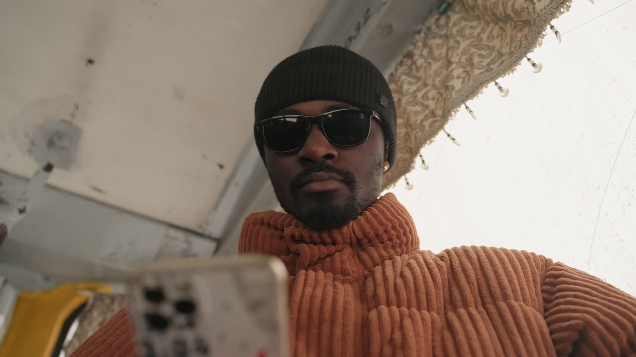 Black man in beanie filming content for social channel, ribbed orange sweater and sunglasses, lowangle closeup inside transit vehicle, confident expression, city backdrop visible, smartphone used