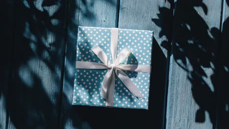 a blue and white polka dot gift box with a white ribbon and bow, sitting on a wooden surface with shadows from leaves