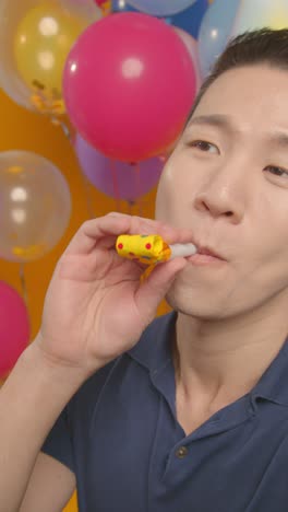 vertical video studio portrait of man wearing party hat celebrating birthday with balloons and party blower 1