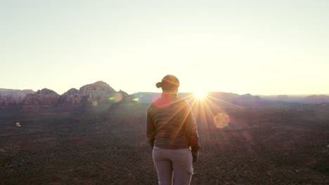 Hiker-stands-on-cliff-at-sunrise-watches-sun-crest-over-mountain-tops-in-Sedona