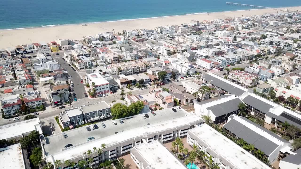 Drone pushes in over a coastal neighborhood in Redondo Beach then tilts up to reveal wide sandy beach ocean and distant pier. Clean rooftops palm lined streets and apartment complexes