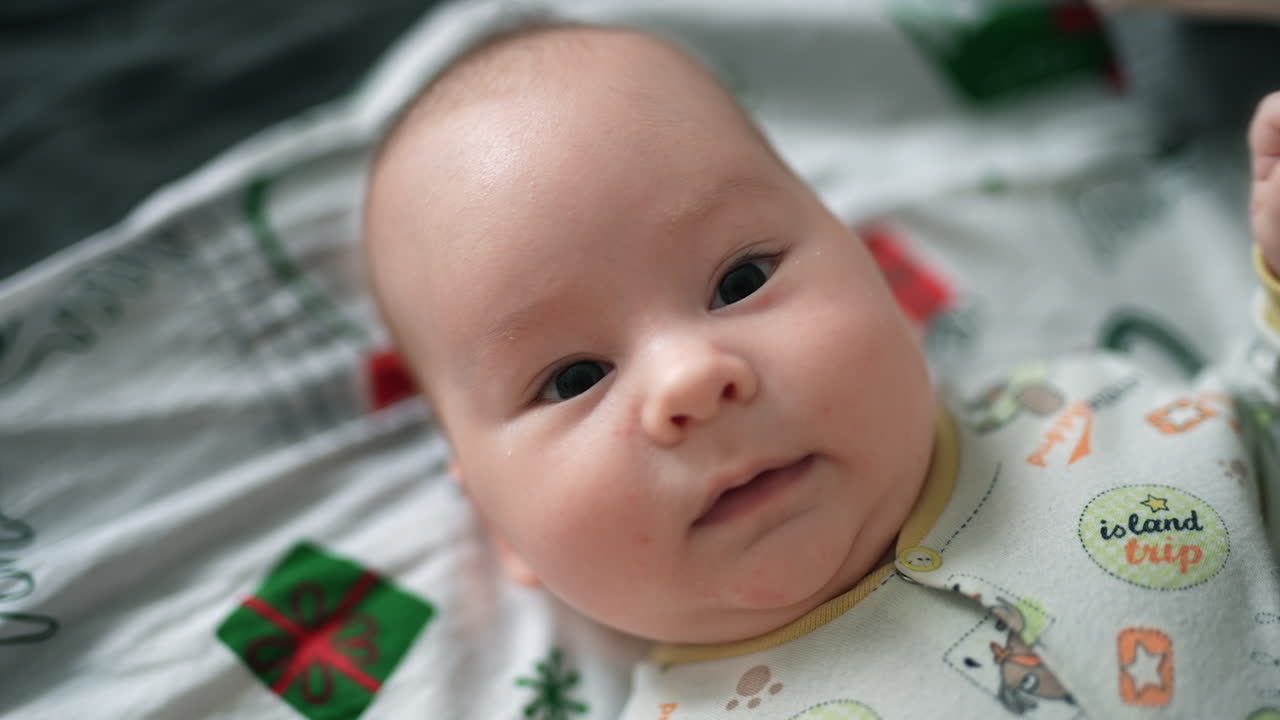 Small infant boy with plump cheeks resting on the bed. Beautiful toddler watching intensely the camera. Top view close up.