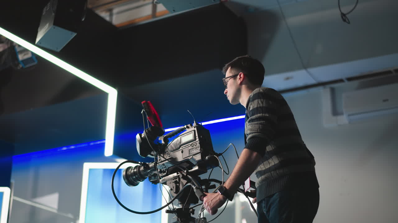 Camera operator adjusting equipment while filming in modern studio. Professional video camera on tripod, surrounded by lighting setups and cables. Focused on technical aspects of video production