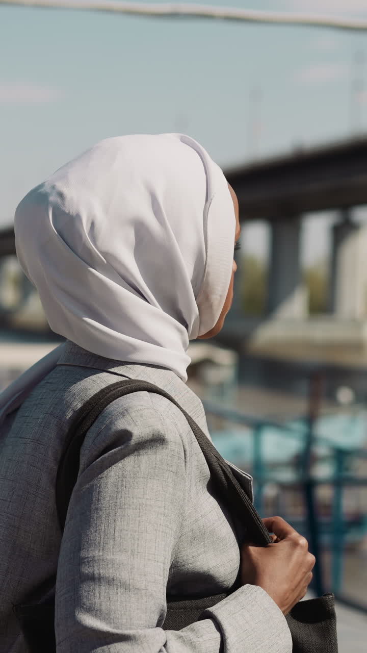 Positive Muslim woman with hijab walks along embankment past barge to bridge slow motion. Black lady on waterfront. Happy young Muslimah in city at sunlight