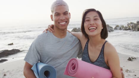 portrait of happy biracial couple holding yoga mats at beach, in slow motion