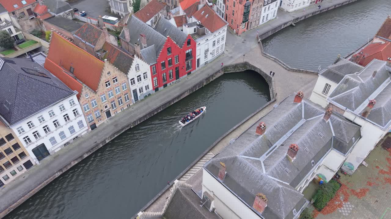 High angle drone shot looking down at a boat full of tourists navigating the historic canals of Bruges, Belgium, and passing under an ancient King's Bridge