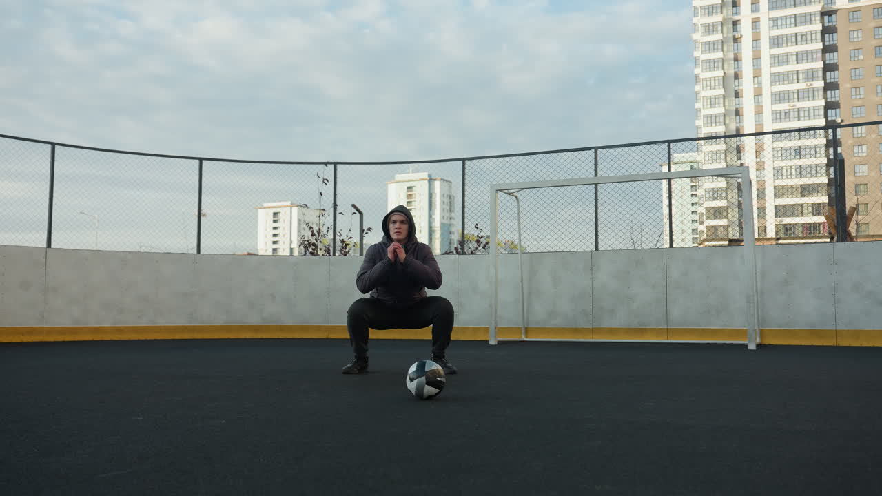 atleta realizando sentadillas con las manos juntas en preparación para el ejercicio en un campo deportivo al aire libre, una pelota de fútbol en primer plano, rodeado de oficinas urbanas de gran altura y postes de gol