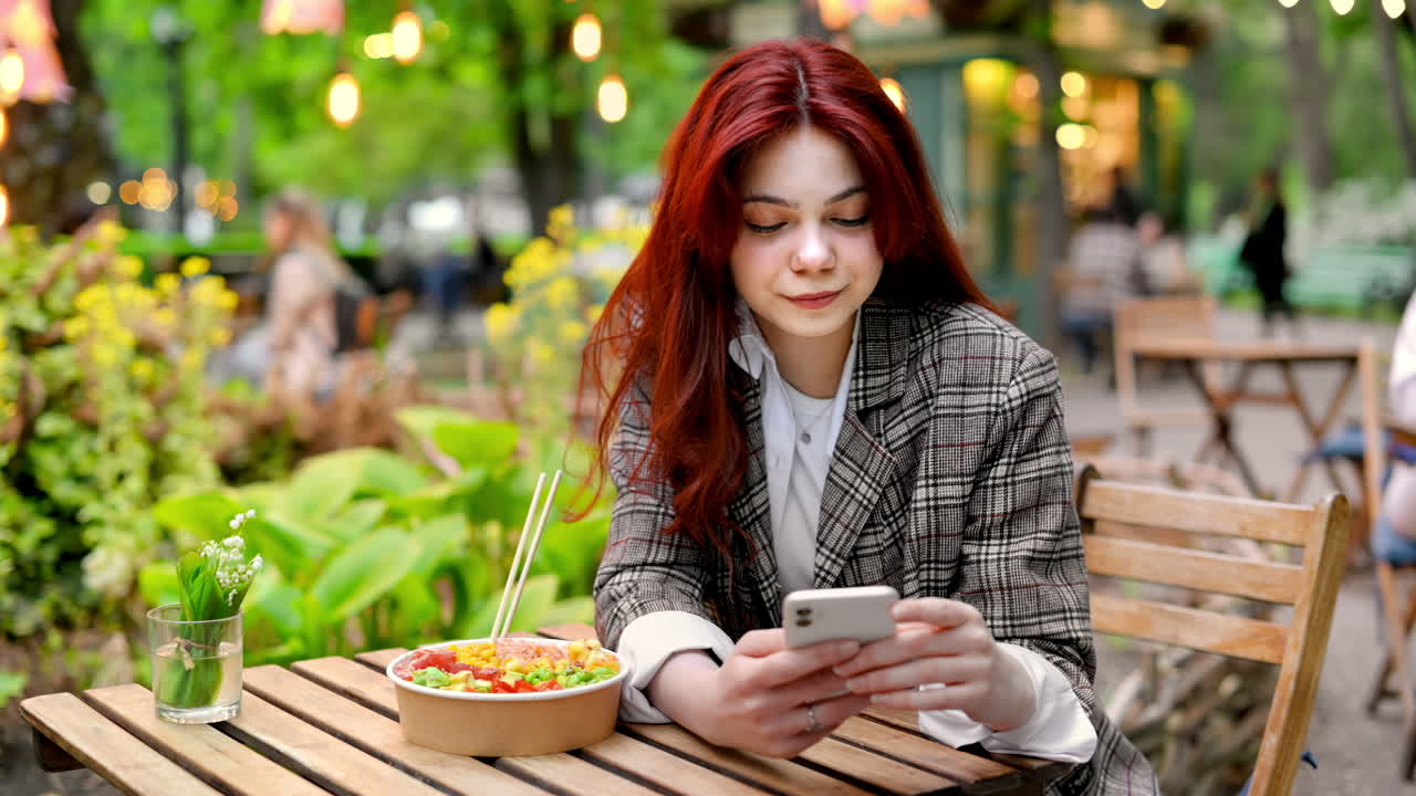 View of a young woman using her phone while sitting on a table with a poke bowl and chopsticks. Park with a lot of greenery and illumination. Slow motion