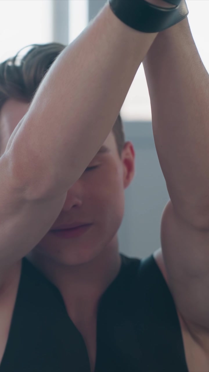 Primo piano di un giovane uomo con una canotta nera che sistema le fasce per i polsi. La corporatura muscolosa è visibile mentre l'uomo fissa l'equipaggiamento tattico. L'illuminazione soffusa enfatizza i dettagli della parte superiore del corpo.