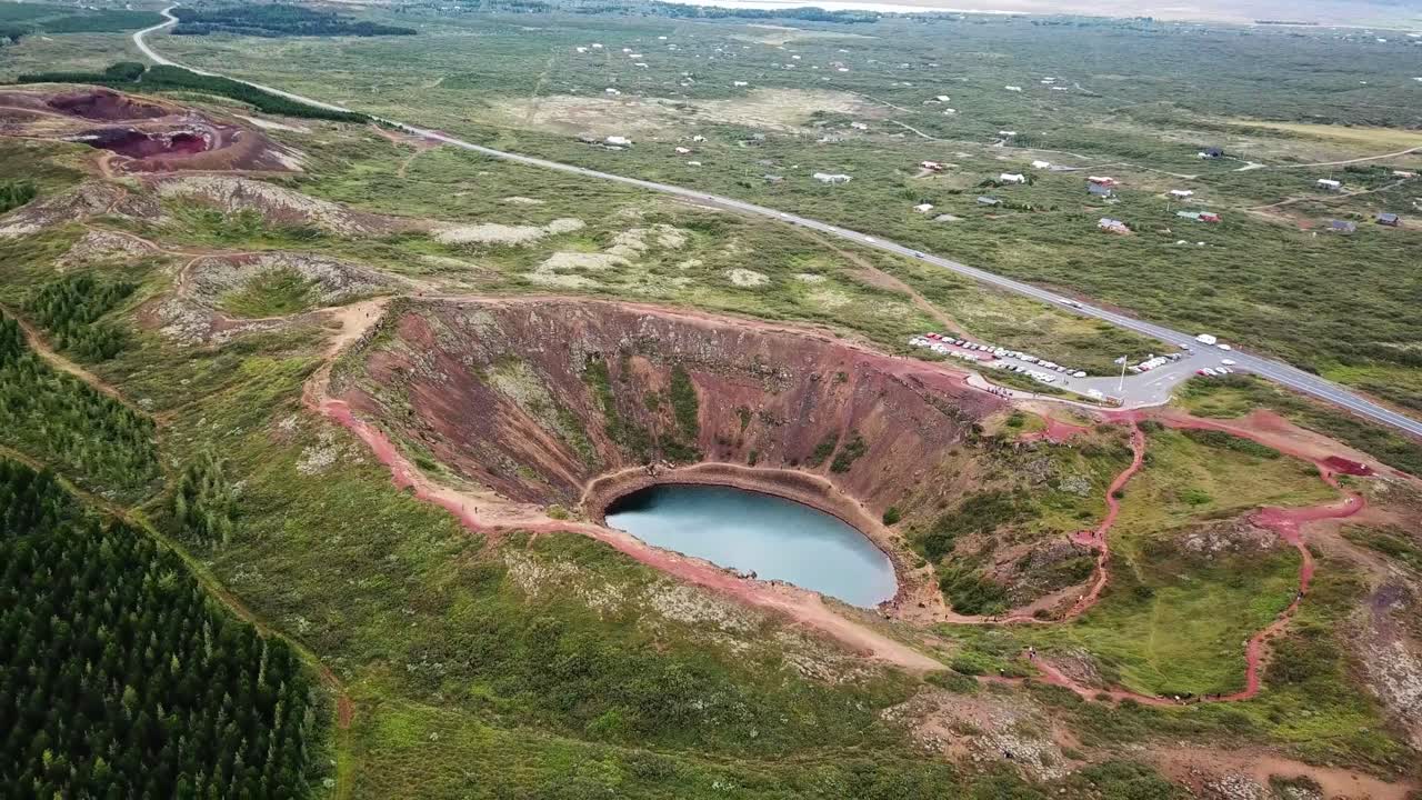 Kerid Volcanic crater lake reflecting blue sky in Iceland Golden Circle, featuring red slopes, green moss and hiking paths, aerial view from above, drone pulling out
