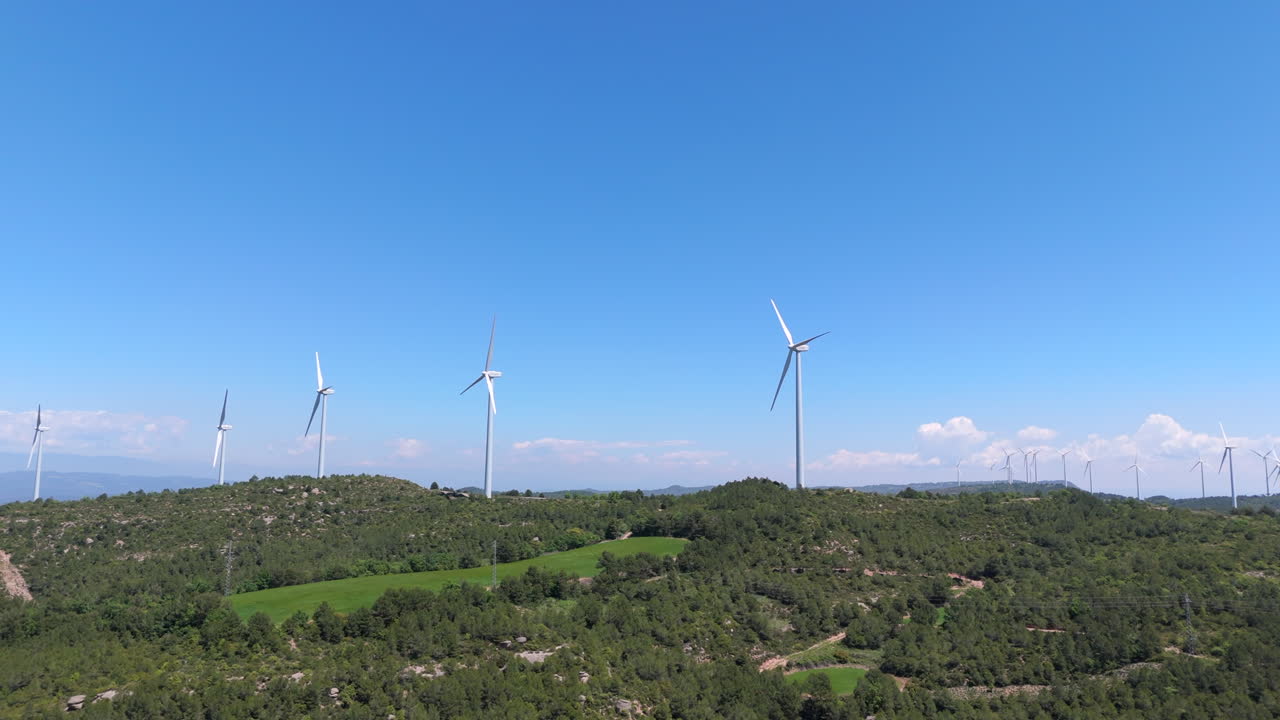 Lateral drone shot of multiple wind turbines spinning on forested hills. Green terrain contrasts with clear blue sky and scattered clouds, emphasizing clean energy and rural elevation
